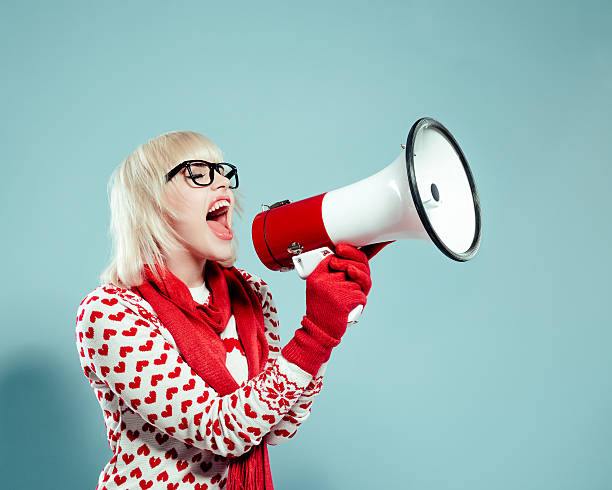 Blonde young woman wearing xmas sweater, screaming into megaphone Portrait of excited blonde young woman wearing christmas sweater, red scarf and gloves, screaming into megaphone. Studio shot, turquoise background. christmas nerd sweater cardigan stock pictures, royalty-free photos & images