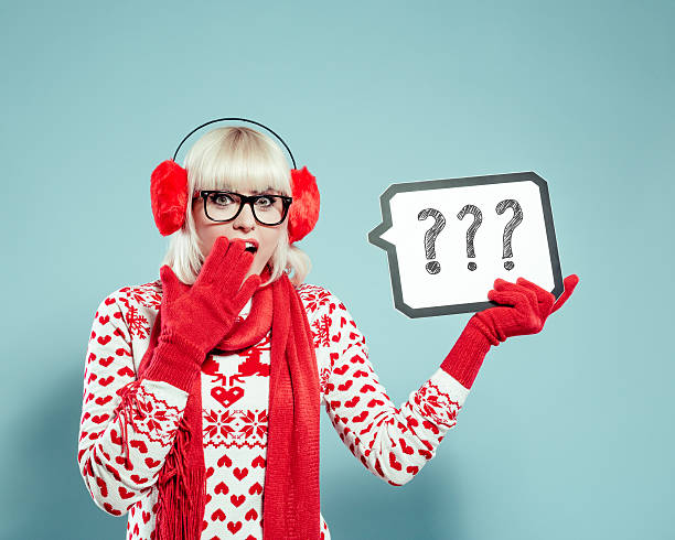 Surprised blonde young woman in winter outfit holding speech bubble Portrait of surprised blonde young woman wearing christmas sweater, red scarf and gloves, nerd glasses and earmuffs, holding speech bubble with drawn question marks, covering her mouth with hand. Studio shot, turquoise background. christmas nerd sweater cardigan stock pictures, royalty-free photos & images