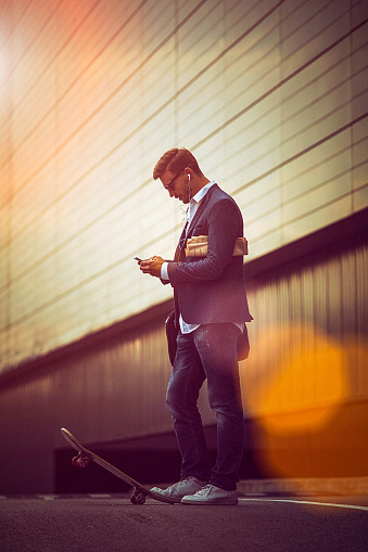 Young businessman using smartphone in the urban environment The man is casually dressed and wears sunglasses, earphones and carries black briefcase hung on shoulder.