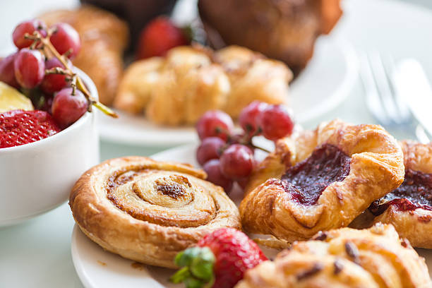 Continental Breakfast - Cinnamon Bun, Danishes, Rolls, Muffins, Fresh Fruit Closeup of a continental breakfast at a hotel, with bowls and plates filled with a selection of breakfast bakery items and fresh fruit on a white table with cutlery in the background. continental breakfast photos stock pictures, royalty-free photos & images
