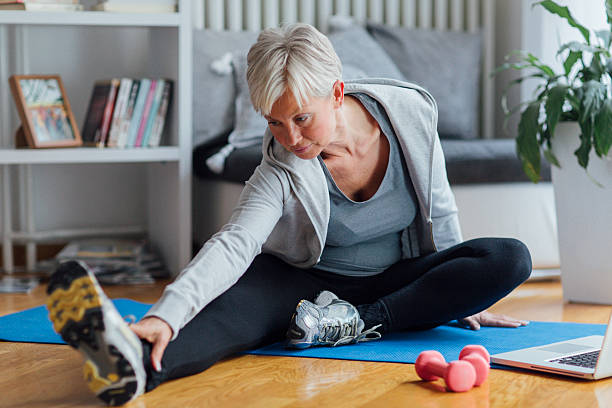 Mature Woman Exercising At Home. Mature woman exercising at home, learning exercises online with laptop. Sitting on floor and stretching. grey hair on floor stock pictures, royalty-free photos & images