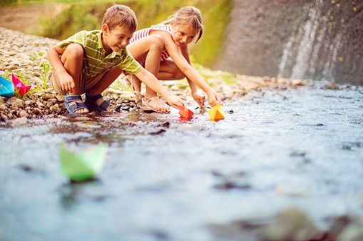 Little boy and girl playing paper boats on the creek. They are very playful and happy while competing in boat race.