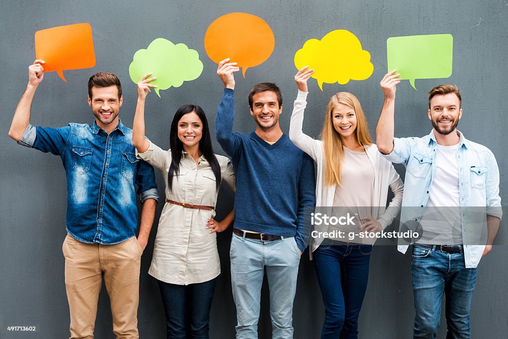 Global communications. Group of happy young people holding empty speech bubbles and looking at camera while standing against grey background Holding Stock Photo