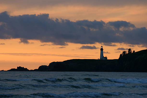 A distant lighthouse on a dark jetty cliff is sihouetted by a a sunset on the Pacific Ocean in America