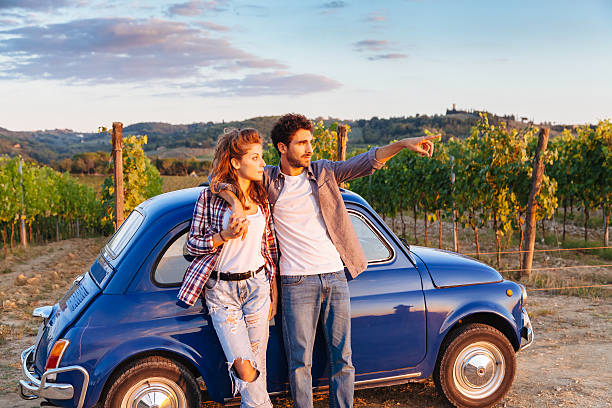 Loving couple watching the sunset A loving couple watch the sunset, standing leaning against an old blue car, during their journey through the countryside of Tuscany, Italy. Around them, rows of vines and the classic hills. The young man shows to his girlfriend sunset time period stock pictures, royalty-free photos & images