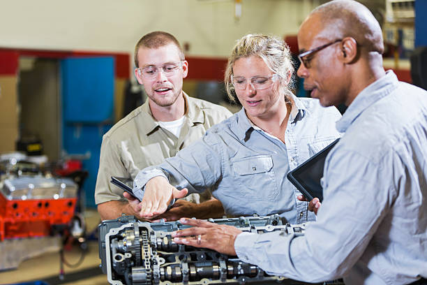 Multi-racial mechanics working on gasoline engine A group of three mult-ethnic mechanics working on a gasoline engine, discussing what needs to be done to repair it.  The mature, African American man in the foreground is out of focus.  The woman standing in the middle is pointing to the engine and the young man standing behind her is watching and holding a digital tablet. multiengine stock pictures, royalty-free photos & images