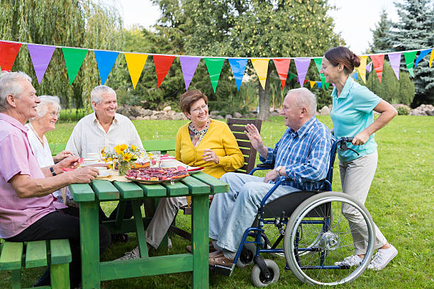 Group of elderly people sitting at the garden table Group of elderly people sitting at the garden table and celebrating birthday with birthday cake. In the foreground a caregiver with an senior man sitting on wheelchair. senior adult women park bench 70s stock pictures, royalty-free photos & images