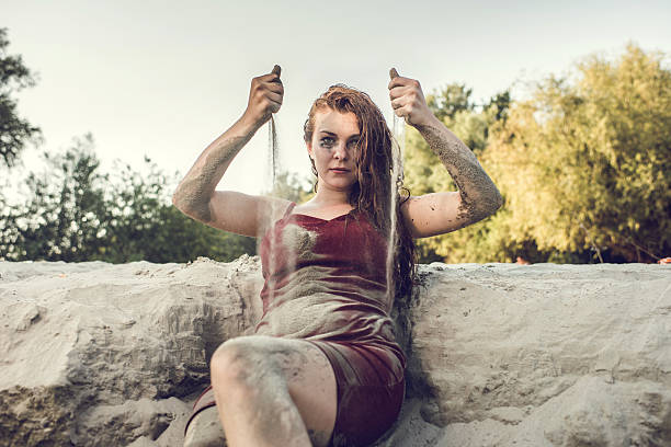 Mentally ill redhead woman sitting on a sand and pouring it from her hands while looking at the camera.
