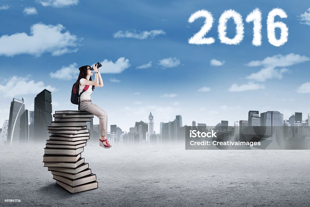 Student on the pile of books looking at numbers 2016 Image of female high school student sitting on the stack of books while using binoculars to see cloud shaped numbers 2016 2015 Stock Photo