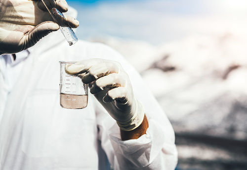 Environmental inspector in white safety suit examing polluted water at industrial site. Heaps of coal covered with white ash in the background.