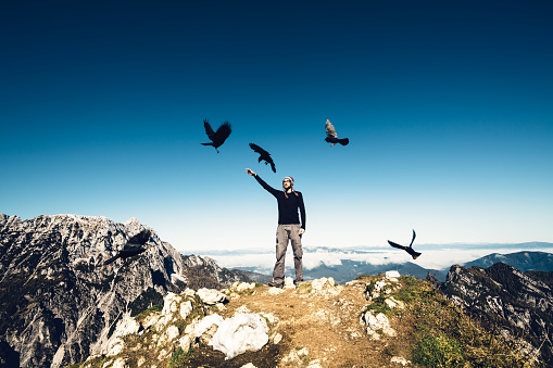 Young man standing on top of the mountain surrounded by black birds. Man is wearing winter hat, black shirt and brown trousers. His arm is outstrached and birds are flying around.