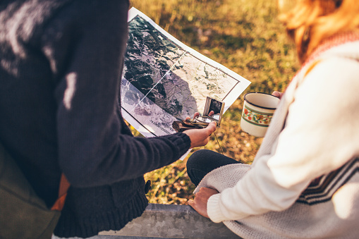 Young couple is using a compass and maps for orientation in the wilds, while resting by the road and drinking tea from the pot