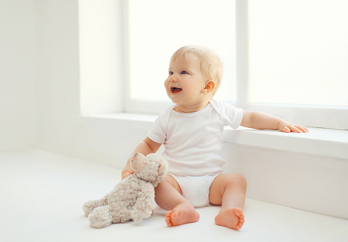 Cute smiling baby with teddy bear toy sitting at home in white room near window
