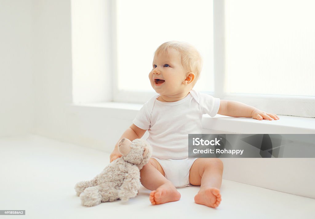 Cute smiling baby with teddy bear toy home in room Cute smiling baby with teddy bear toy sitting at home in white room near window Baby - Human Age Stock Photo