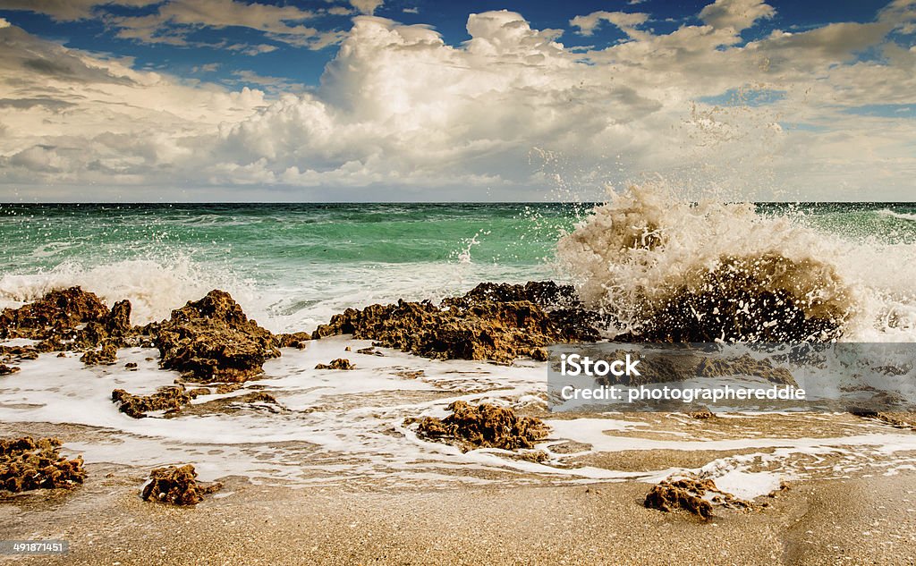 Waves Crashing Against the Rocks This photograph depicts a cloudy blue sky behind a greenish colored ocean with rough waves crashing in at Hutchinson Island.  Hutchinson Island is a barrier island on the coast of Martin and St. Lucie counties, Florida. The southern one-third of Hutchinson Island is in Martin County while the northern two-thirds is in St. Lucie County. It is bounded on the east by the Atlantic Ocean, on the south by the St. Lucie Inlet, on the west by the Indian River, and on the north by the Fort Pierce Inlet, which separates it from what is known in St Lucie County as North Hutchinson Island, or sometimes "North Beach." North Hutchinson Island extends into Indian River County, but it is rarely called that north of the county line. In fact the Indian River county portion which extends to the Sebastian Inlet is usually called Orchid Island, although it is not a separate island. Florida - US State Stock Photo