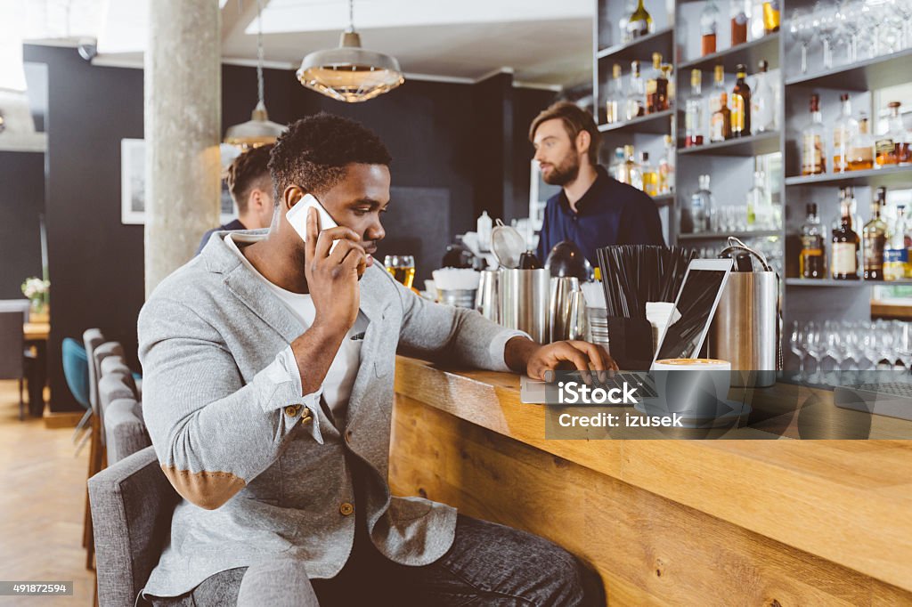 Afro american man using laptop in a pub Afro american man wearing jacket sitting by the counter in the pub, using laptop and talking on mobile phone. Bearded barman in the background.  Laptop Stock Photo