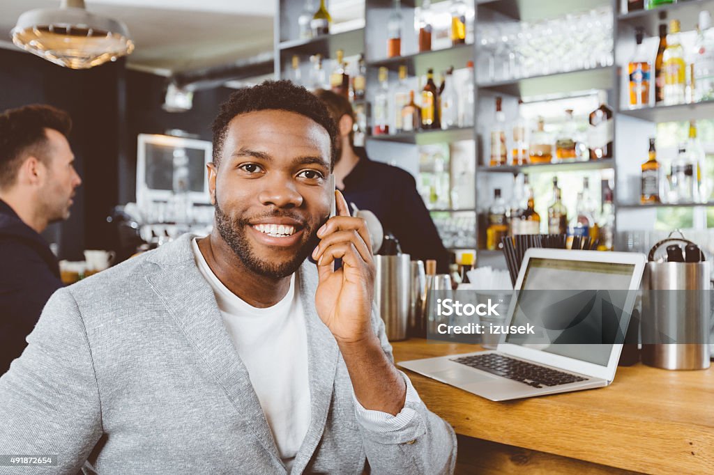 Afro american man talking on phone in the pub Afro american man wearing jacket sitting by the counter in the pub, talking on mobile phone. Laptop on the counter. 2015 Stock Photo