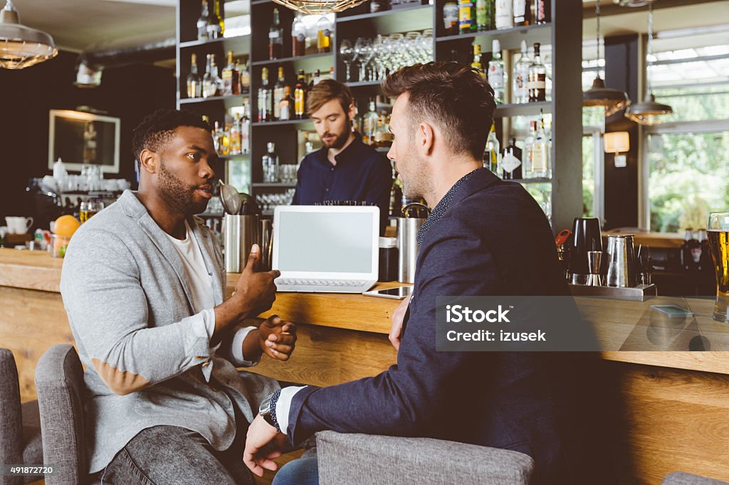 Afro american man talking with colleague in pub, using laptop Afro american man wearing jacket sitting by the counter in the pub and talking with caucasian businessman. Laptop on the bar counter, barman in the background.  Bar - Drink Establishment Stock Photo