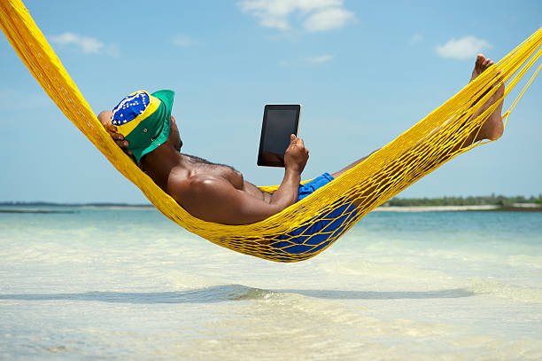 Brazilian Man Relaxing with Tablet in Beach Hammock Brazilian man using tablet computer relaxing in hammock on beach over the sea hammock men lying down digital tablet stock pictures, royalty-free photos & images