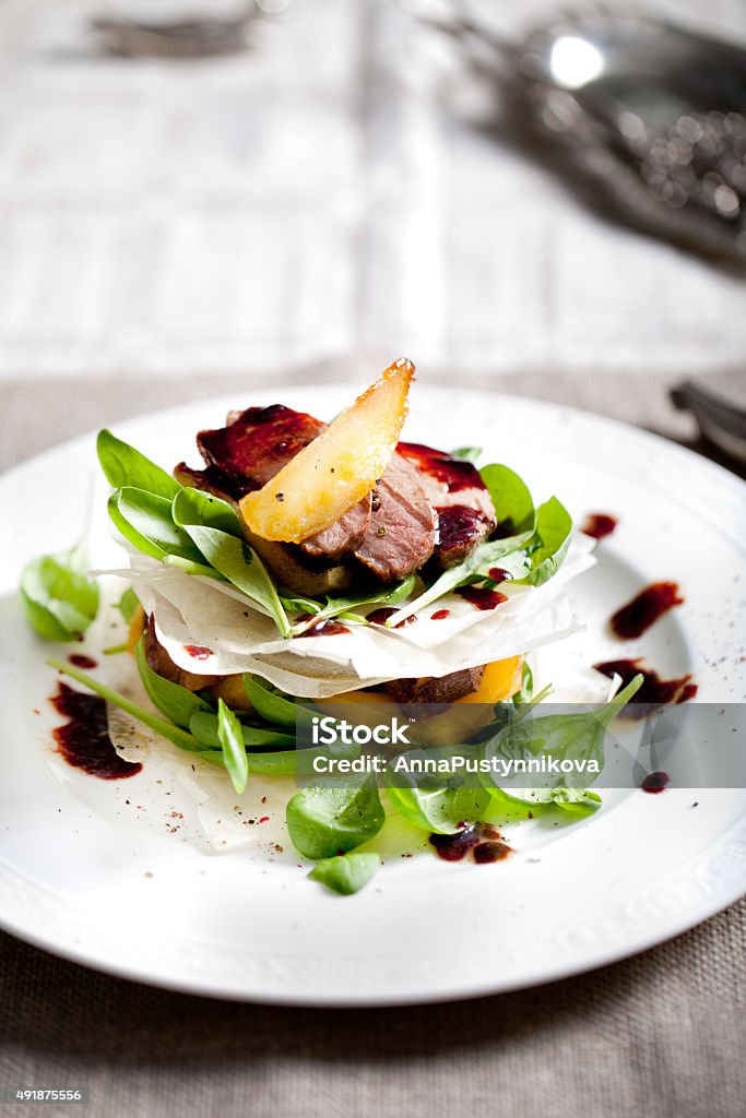 Duck breast roasted with salad, pear Duck breast roasted with caramelized pear, baby spinach and crispy filo pastry sheets with berry sauce on a white plate on a textile background Gourmet Stock Photo