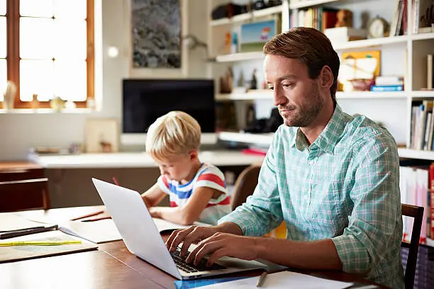 Father working on laptop and son coloring at table in living room