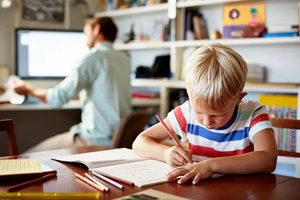 Boy coloring at table with his dad working in the background