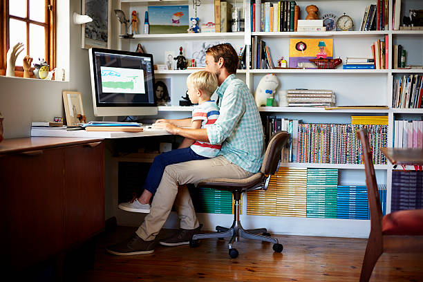 Boy sitting on father's lap at computer desk Boy sitting on father's lap at computer desk in living room home office chair stock pictures, royalty-free photos & images