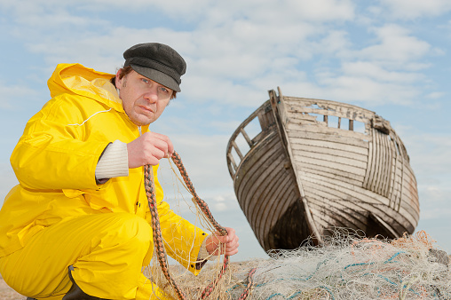 Fisherman wearing yellow water-proof overalls whilst repairing his nets, with an old wooden fishing boat in the background.