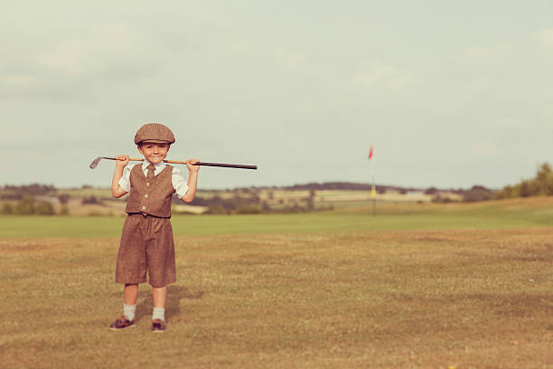 A little boy dressed in vintage golfing attire stands with his club on a golf course in England. He is standing by the putting green and is smiling because he loves golf.