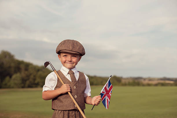 A little boy dressed in vintage golfing attire stands with his club on a golf course in England while he is holding the flag of the United Kingdom. He is wearing a vest and tie with a vintage hat. He is smiling because he loves golf.