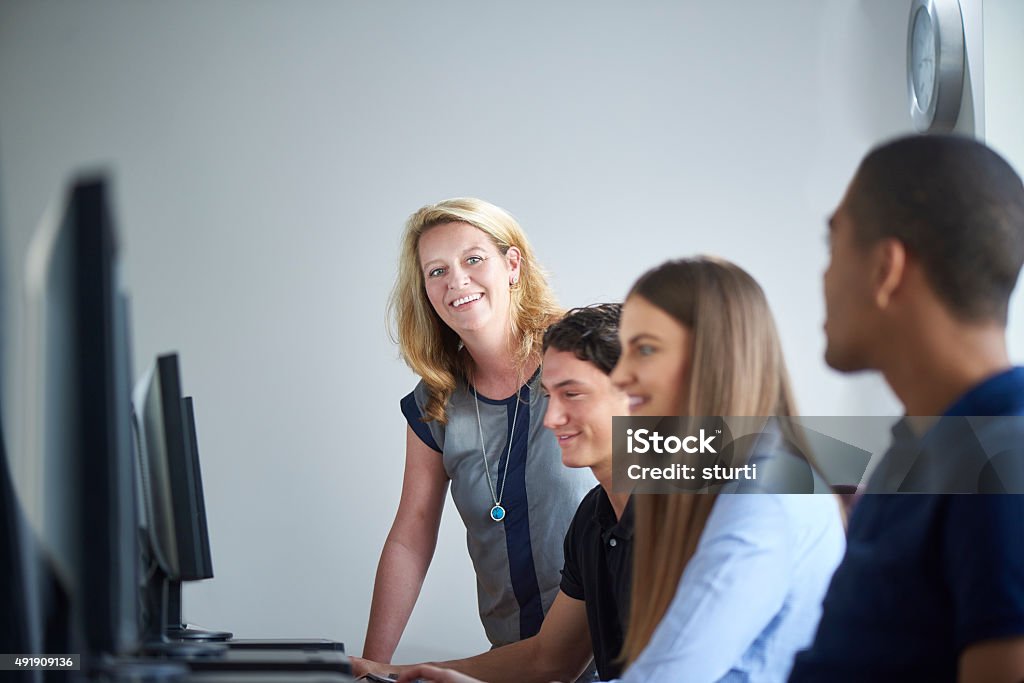 computer room teacher female teacher chats to students in the computer lab . She is standing and looking along the row of students . Teacher Stock Photo