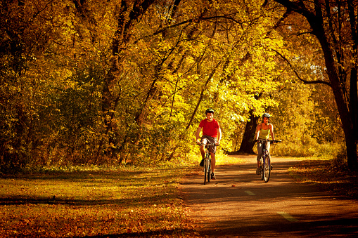Two bicyclists bicycling on a park bike path in Autumn color. Young Caucasian man and a black American young woman exercising together in a nature park together. Photographed on location in Minneapolis,f Minnesota, USA in horizontal format with available copy space.
