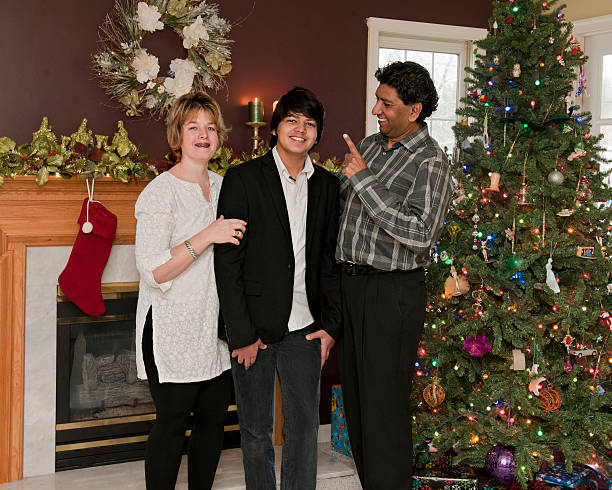 A family of 3 standing in their Christmas-decorated living room.  The dad is teasingly warning his young teen son while the boy and wife pose for a portrait.