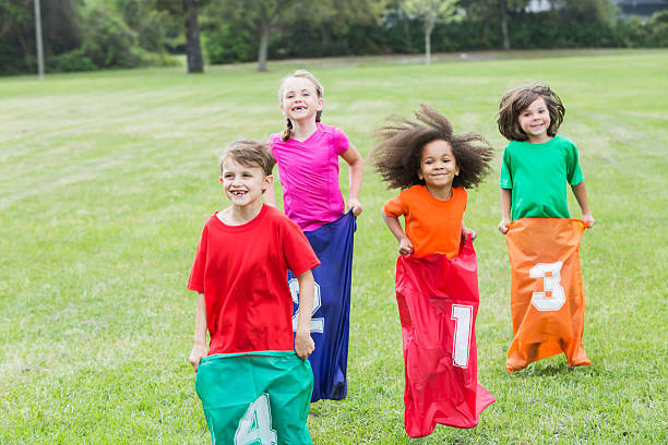 A multiracial group of four children in a potato sack race in the park.  They are wearing colorful shirts and jumping in colorful, numbered sacks.