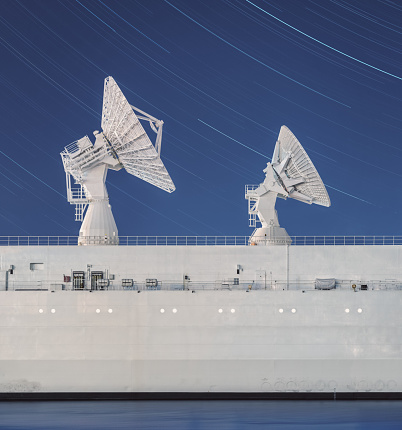 The stars are set into motion above two satellite dishes perched high atop the deck of a vessel used in tracking rocket trajectories.  Long exposure.