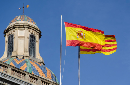 Two flags waving in the blue sky : Spanish and Catalan, and an old building dome beside, Picture taken in the streets of Barcelona, Spain