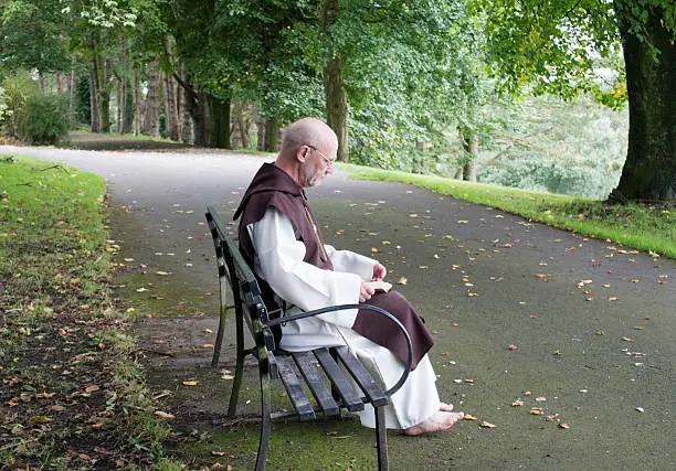 Mature monk sitting on a park bench reading a Bible outdoors