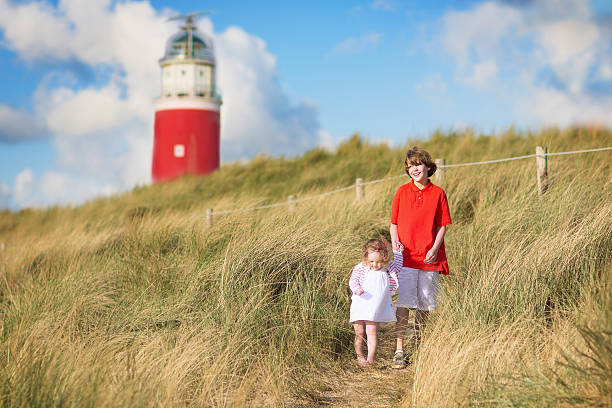 Happy children, young active boy and his adorable curly baby sister wearing a dress playing with sand toys on a sunny windy beach with a red lighthouse on Texel island, Holland, Netherlands