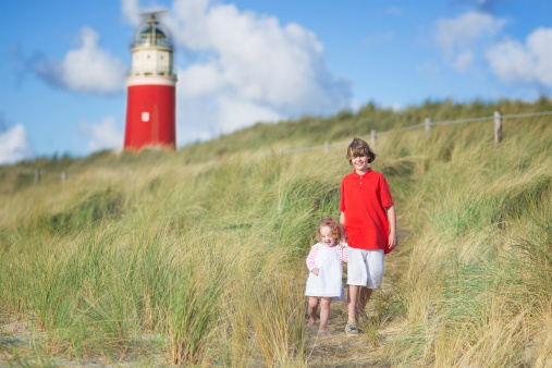 Happy children, young active boy and his adorable curly baby sister wearing a dress playing with sand toys on a sunny windy beach with a red lighthouse on Texel island, Holland, Netherlands