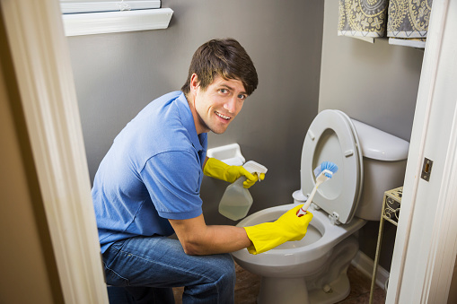 A man doing housework.  He is kneeling in the bathroom at the toilet bowl, wearing rubber gloves, with a spray bottle and scrub brush.  He is looking at the camera and smiling.