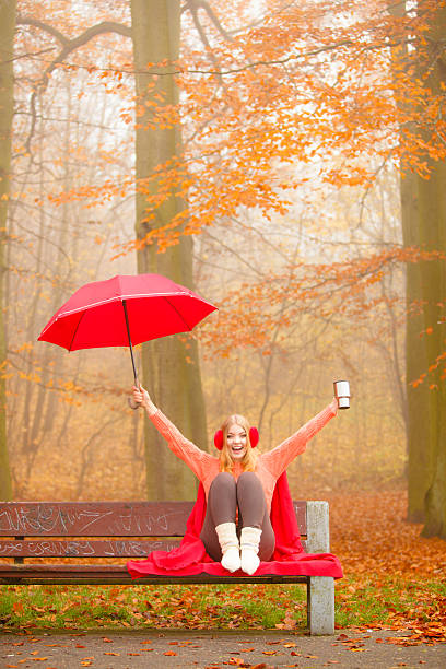 Happiness carefree and fall concept. Young woman in red relaxing in autumn park on bench under umbrella, enjoying hot drink holding mug with warm beverage. Orange leaves background