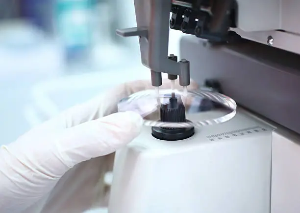 Closeup of unrecognizable caucasian technician's hand placing piece of lens glass into machine that will mark the focal center before sending it to glass grinder. The technician is wearing white protective gloves.