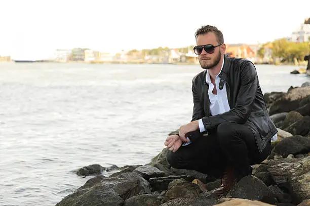 An attractive man in his 20s wearing a white shirt and black leather jacket with dark sunglasses, being by the water in a harbor. 