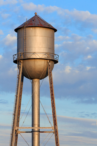 An old metal water tank, in the light of the morning sun, stands tall against a cloudy blue sky in America's Midwest.