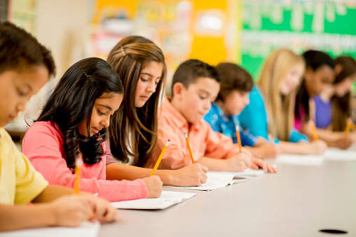 A multi-ethnic group of elementary age children are sitting in a row in class and are taking exams.