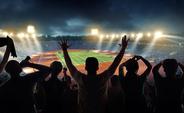 Fans at . stadium with running tracks On the foreground a group of cheering fans watch a sport championship on stadium. One man stands with his hands up to the sky. People are dressed in bright colors. A long-range shot of a stadium field, floodlights and seating. A green field, with painted white lines and running tracks is visible in the foreground. In the background are diffuse out-of-focus stadium seats. Large, bright floodlights are in the top-left and top-right corners of the image. bleachers photos stock pictures, royalty-free photos & images