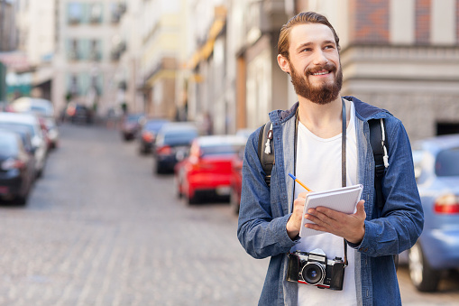 Cheerful male tourist is standing with camera and backpack in town. He is looking at architecture with interest and writing some notes. The man is smiling. Copy space in left side