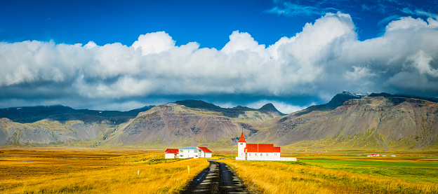 White-washed church, rural village homes and farmhouses with characteristic bright red roofs surrounded by pastoral plains beneath the dramatic cloud topped mountain ridges of the Snaefellness peninsula of Iceland. ProPhoto RGB profile for maximum color fidelity and gamut.