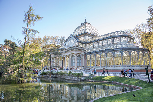 Madrid, Spain - April 5, 2015: Tourists and locals gather outside the Crystal Palace (Palacio de Cristal) in the Retiro Park in Madrid on a cold winter afternoon. Designed by architect Ricardo Velázquez Bosco, it was built in 1887 to exhibit flora and fauna from the Philippines. 
