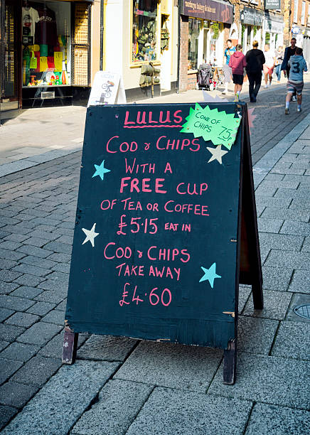 Cod and Chips - sign King's Lynn, Norfolk, England - September 19, 2015: A sign outside a fish and chip shop in Tower Street, King’s Lynn, Norfolk, England, advertising cod and chips. A few people are walking in the bright sunshine further along the shop-lined street. kings lynn stock pictures, royalty-free photos & images